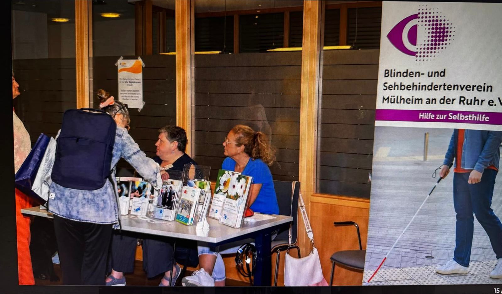Ein Informationsstand des Blinden- und Sehbehindertenvereins Mülheim an der Ruhr e. V. ist aufgebaut. Hinter dem Tisch sitzen zwei Frauen: links Maria St. Mont (Vorsitzende) und rechts Nicole Schmitz-Siegfried (Vorstandsmitglied). Auf dem Tisch stehen zahlreiche Flyer, Informationsmaterialien und ein Kalender mit Bildern von Blindenführhunden. Eine Besucherin mit großem Rucksack und Einkaufstasche beugt sich über den Tisch und spricht mit den beiden. Rechts steht ein Roll-Up mit dem Logo des Vereins, dem Schriftzug „Hilfe zur Selbsthilfe“ und einem Bild eines Mannes mit Blindenstock. Ein Informationsstand des Blinden- und Sehbehindertenvereins Mülheim an der Ruhr e. V. ist aufgebaut. Hinter dem Tisch sitzen zwei Frauen: links Maria St. Mont (Vorsitzende) und rechts Nicole Schmitz-Siegfried (Vorstandsmitglied). Auf dem Tisch stehen zahlreiche Flyer, Informationsmaterialien und ein Kalender mit Bildern von Blindenführhunden. Eine Besucherin mit großem Rucksack und Einkaufstasche beugt sich über den Tisch und spricht mit den beiden. Rechts steht ein Roll-Up mit dem Logo des Vereins, dem Schriftzug „Hilfe zur Selbsthilfe“ und einem Bild eines Mannes mit Blindenstock.