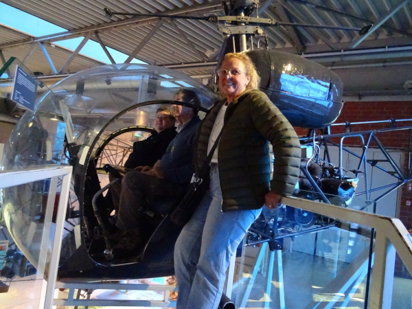 Ein älterer Hubschrauber im Ausstellungsraum, vermutlich ein Museumsstück. Drei Personen sind zu sehen: Zwei sitzen in der Kabine des Hubschraubers, eine Frau steht vorne an der Einstiegstreppe und lächelt. Die Kabine des Hubschraubers ist durch eine große, runde Glaskuppel deutlich sichtbar. Im Hintergrund sieht man moderne Gebäudestrukturen mit Metallträgern und Lampen. Die Atmosphäre wirkt freundlich und interessiert, vermutlich ein Museumsbesuch. Ein älterer Hubschrauber im Ausstellungsraum, vermutlich ein Museumsstück. Drei Personen sind zu sehen: Zwei sitzen in der Kabine des Hubschraubers, eine Frau steht vorne an der Einstiegstreppe und lächelt. Die Kabine des Hubschraubers ist durch eine große, runde Glaskuppel deutlich sichtbar. Im Hintergrund sieht man moderne Gebäudestrukturen mit Metallträgern und Lampen. Die Atmosphäre wirkt freundlich und interessiert, vermutlich ein Museumsbesuch.