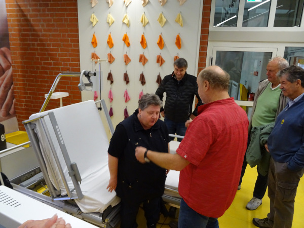 Eine Gruppe erwachsener Männer steht in einem Raum mit gelbem Boden, einer roten Backsteinwand und einer Wand, an der zahlreiche kleine Stoffstücke in verschiedenen Farben hängen. Im Vordergrund ist ein elektrisch verstellbares Pflegebett zu sehen, auf dem eine Person sitzt. Die Gruppe scheint sich auf eine Erklärung oder Demonstration zu konzentrieren, vermutlich im Zusammenhang mit dem Pflegebett oder dessen Funktionen. Die Atmosphäre wirkt wie bei einer Führung oder einem Workshop in einer Einrichtung, die sich mit Pflege oder Medizin beschäftigt. Eine Gruppe erwachsener Männer steht in einem Raum mit gelbem Boden, einer roten Backsteinwand und einer Wand, an der zahlreiche kleine Stoffstücke in verschiedenen Farben hängen. Im Vordergrund ist ein elektrisch verstellbares Pflegebett zu sehen, auf dem eine Person sitzt. Die Gruppe scheint sich auf eine Erklärung oder Demonstration zu konzentrieren, vermutlich im Zusammenhang mit dem Pflegebett oder dessen Funktionen. Die Atmosphäre wirkt wie bei einer Führung oder einem Workshop in einer Einrichtung, die sich mit Pflege oder Medizin beschäftigt.