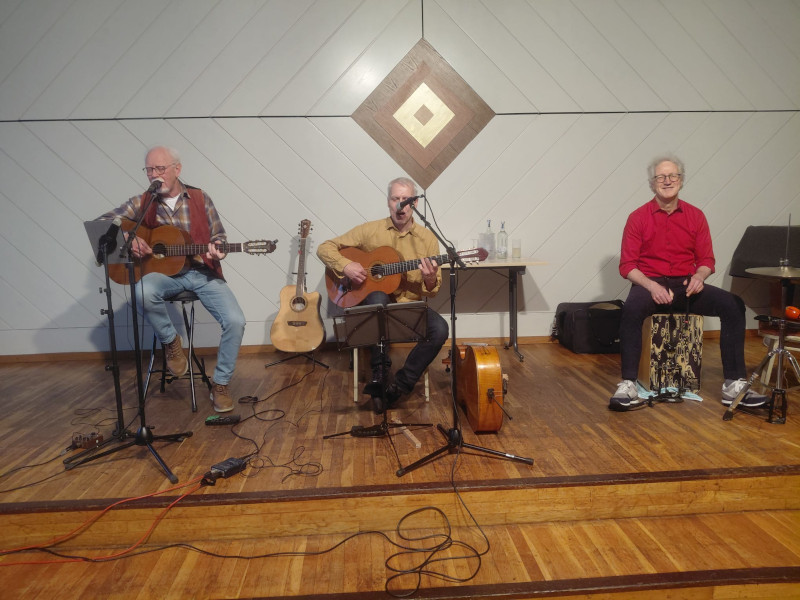 Drei ältere Männer sitzen auf einer Bühne und machen Livemusik. Zwei von ihnen spielen Gitarre und singen, der dritte sitzt auf einer Cajón und lächelt. Im Hintergrund stehen weitere Instrumente und Getränkeflaschen auf einem Tisch. Die Stimmung wirkt entspannt und musikalisch. Drei ältere Männer sitzen auf einer Bühne und machen Livemusik. Zwei von ihnen spielen Gitarre und singen, der dritte sitzt auf einer Cajón und lächelt. Im Hintergrund stehen weitere Instrumente und Getränkeflaschen auf einem Tisch. Die Stimmung wirkt entspannt und musikalisch.
